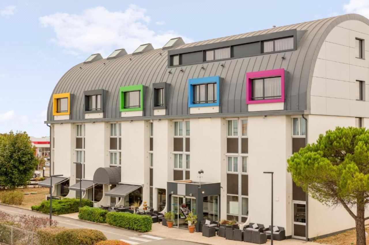 Façade de l'hôtel avec sa toiture arrondie en zinc, ses lucarnes colorées, les stores de la terrasse et le jardin